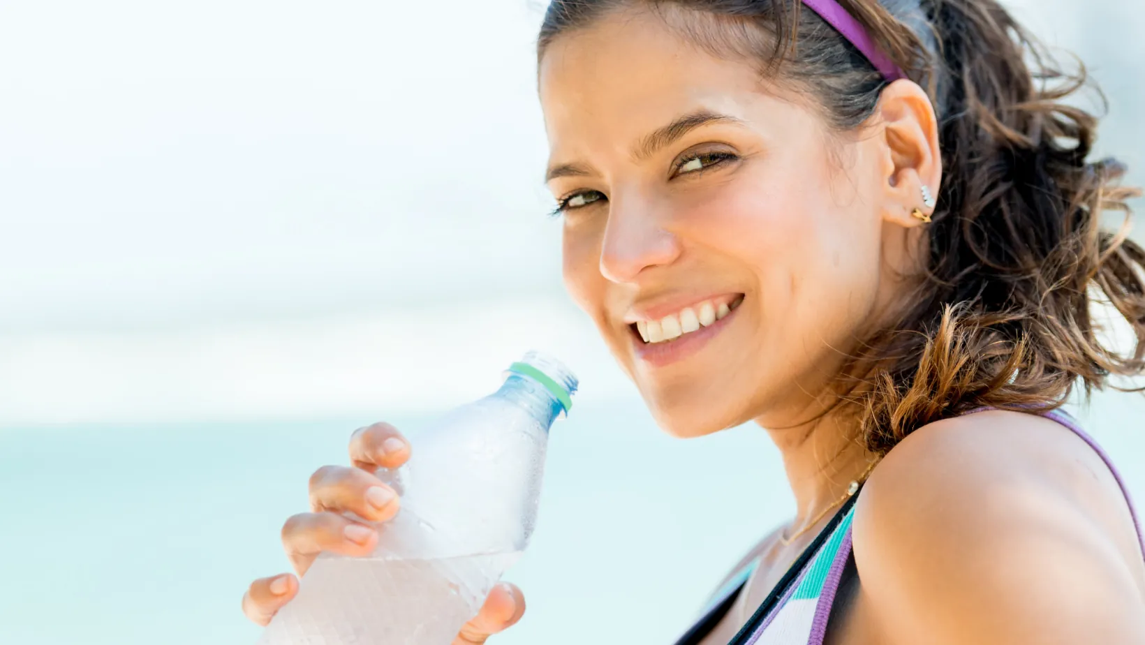 Gewohnheiten zum abgewöhnen: Eine junge sportlich gekleidete Frau trinkt aus einer Flasche Wasser und lächelt in die Kamera